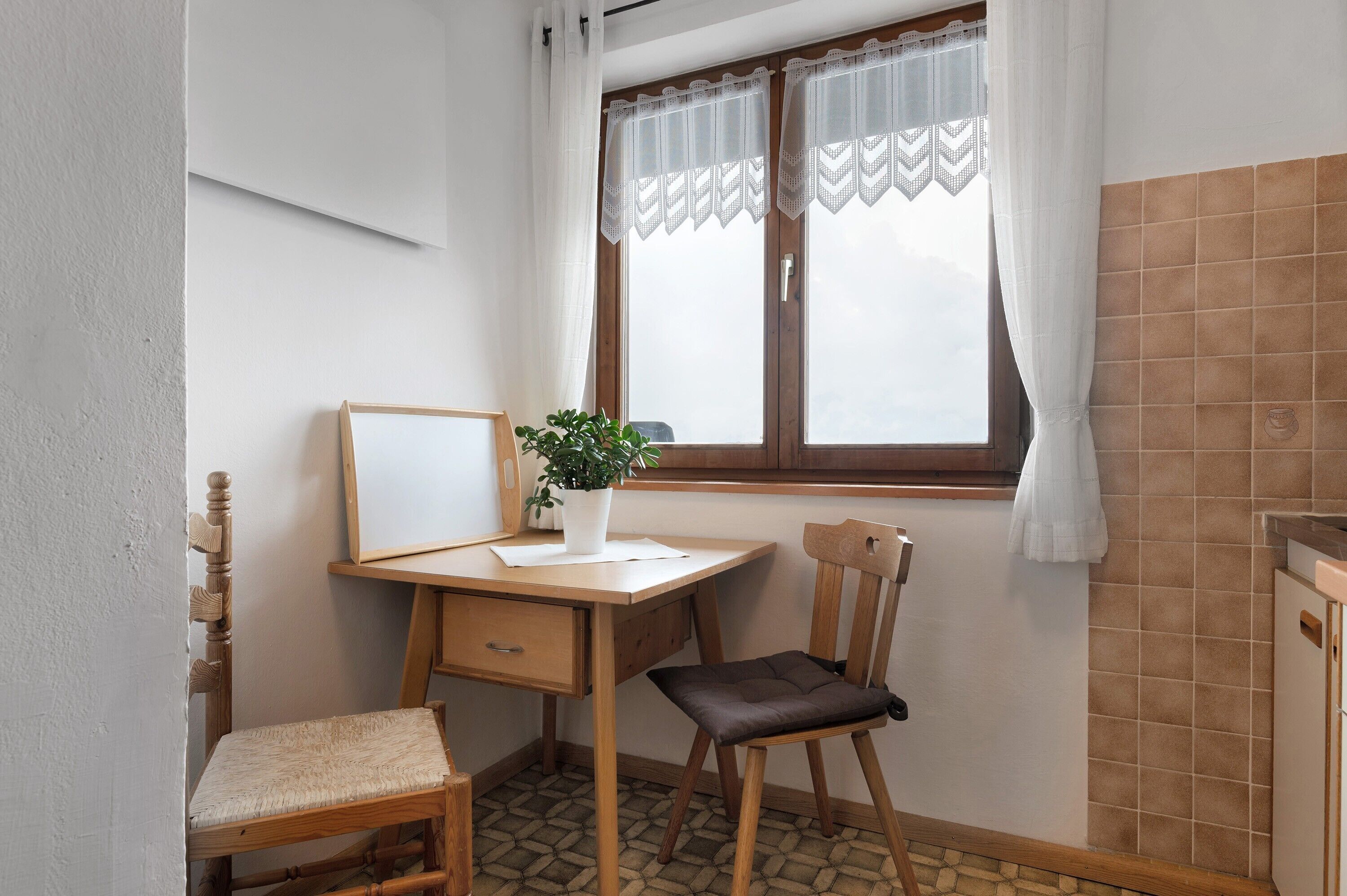 Una habitación pequeña con un escritorio de madera, una silla, una planta en maceta y un ventanal con cortinas.