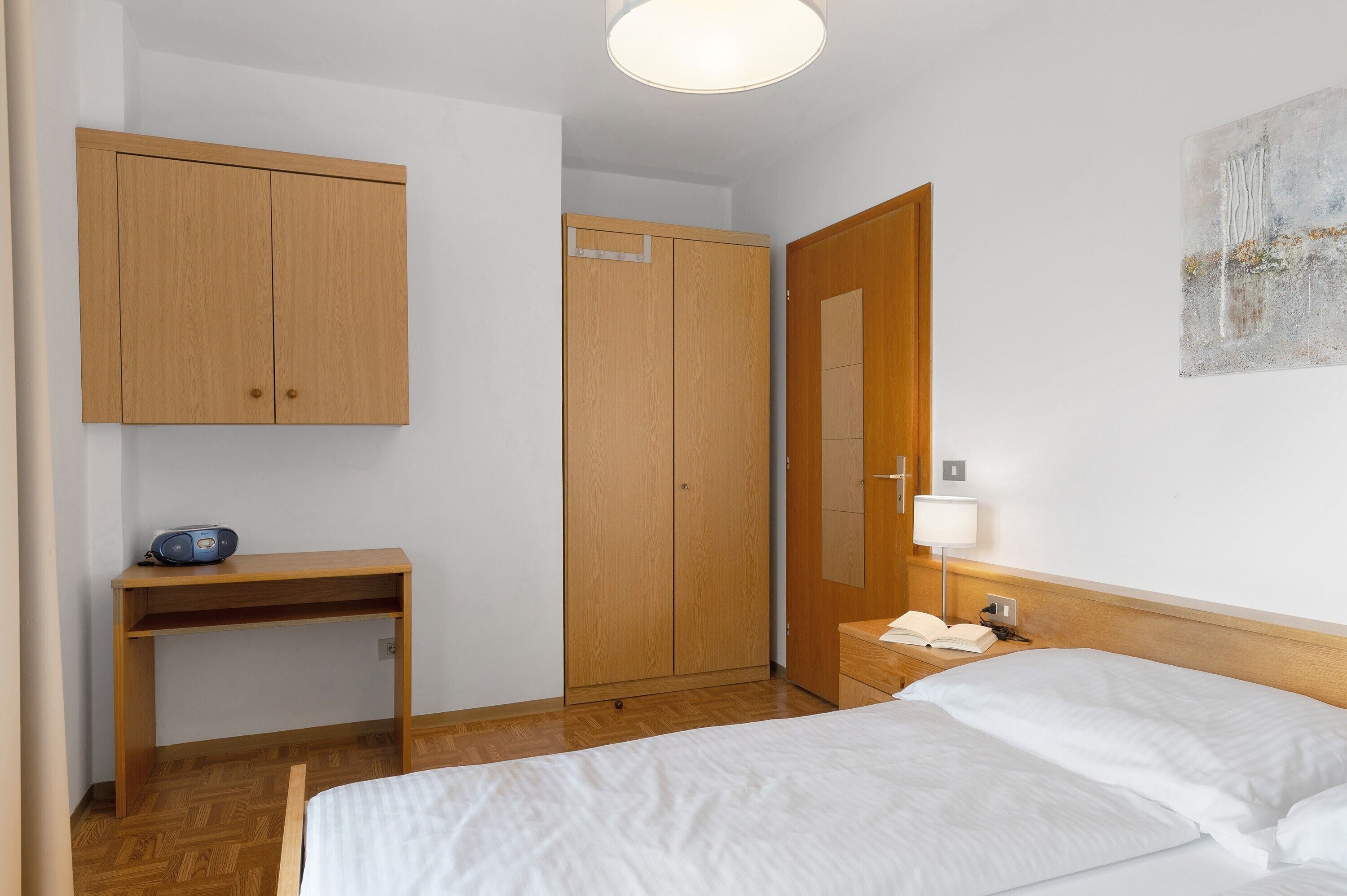 A bedroom with a bed, wooden wardrobe, desk, bedside table, lamp, and a painting on the wall.