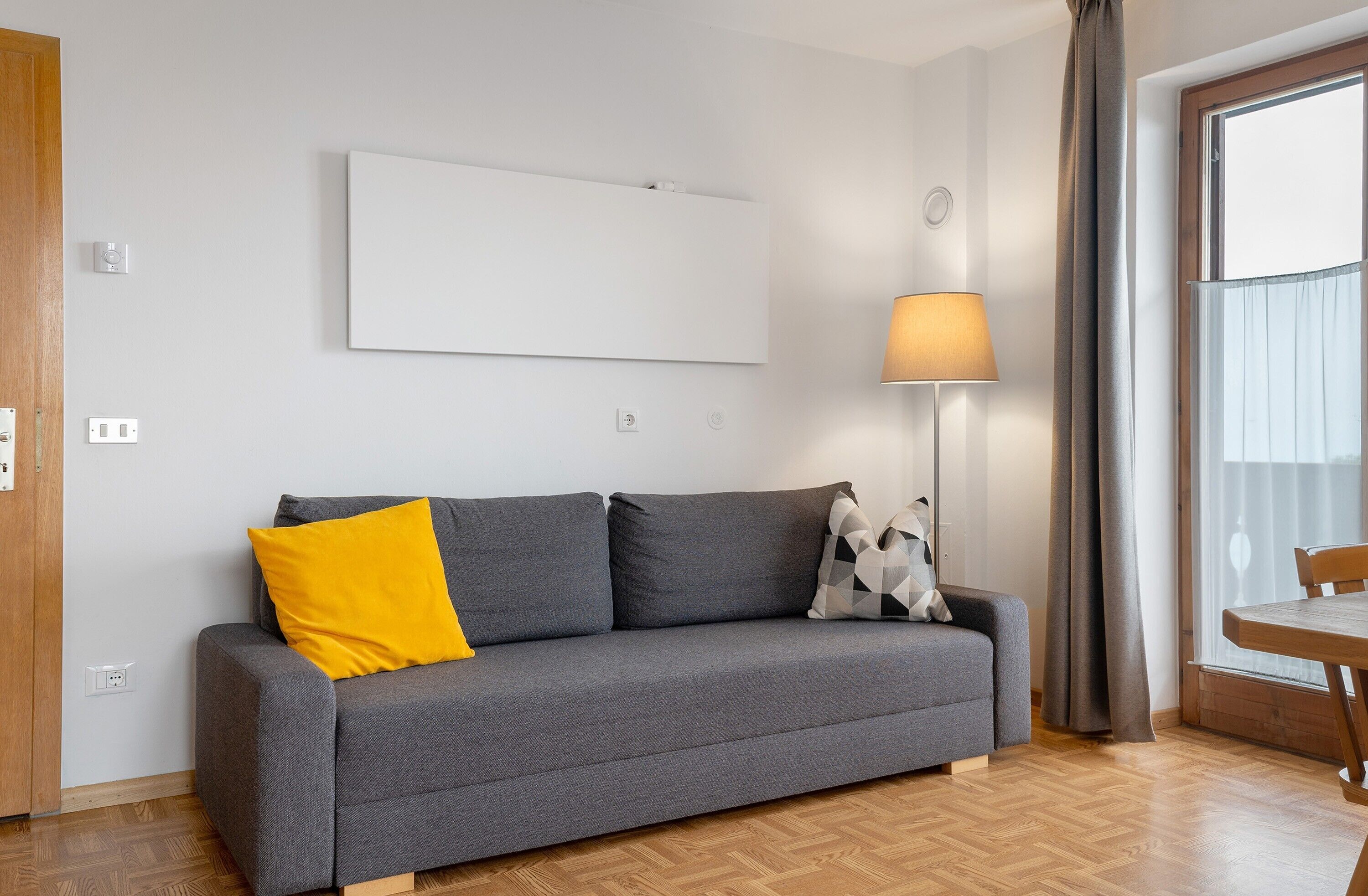 A grey sofa with a yellow pillow, a patterned pillow, a lamp, and a window with curtains.