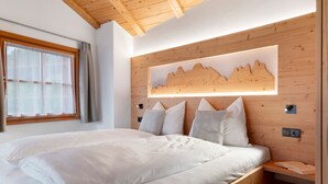 A bedroom with a wooden headboard, a bed with white bedding, a window with curtains, and a wall-mounted light.
