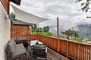 Een houten terras met een tafel en stoelen, een witte zonnescherm en uitzicht op bergen en groen.