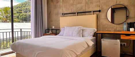 A modern bedroom with a large bed, wooden flooring, a built-in desk, and a view of a lush landscape.