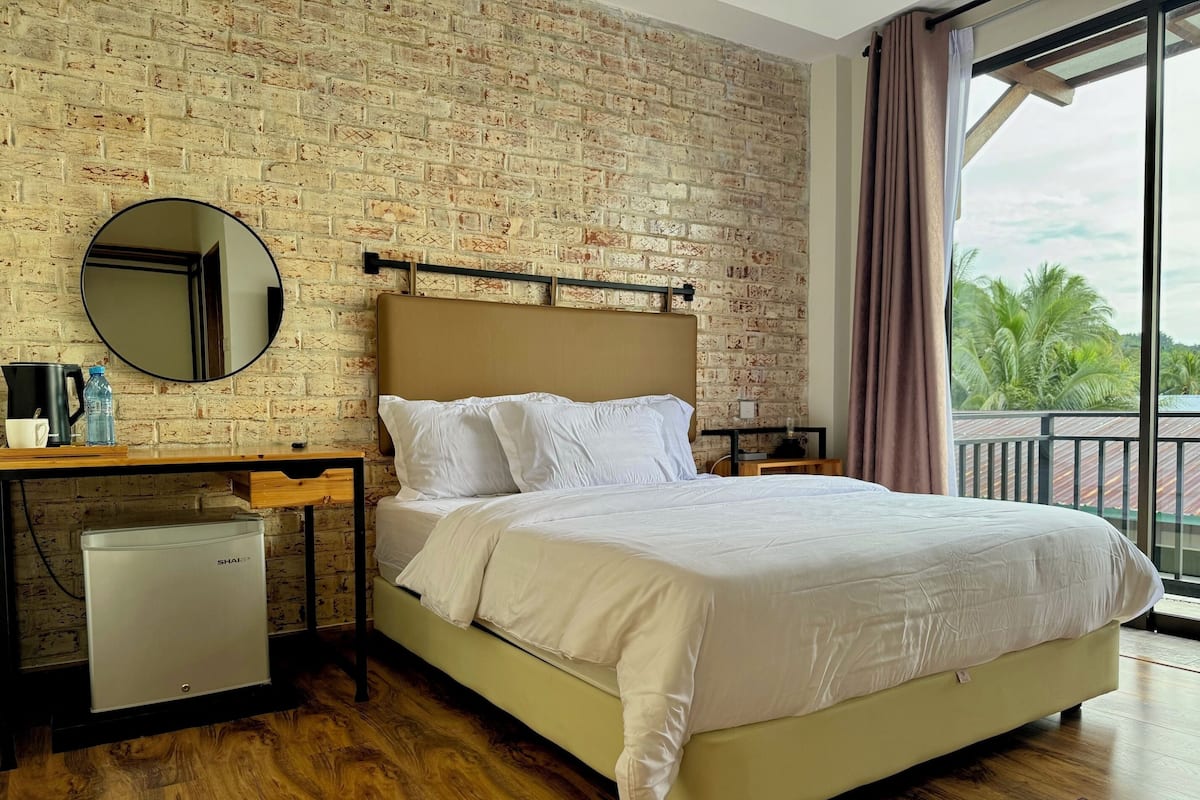 Une chambre d’hôtel avec un mur en briques, un lit avec une literie blanche, un petit réfrigérateur, un miroir rond et une fenêtre donnant sur des palmiers.
