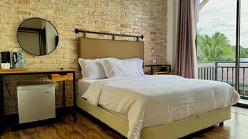 A hotel room with a brick wall, a bed with white bedding, a small refrigerator, a round mirror, and a window with a view of palm trees.
