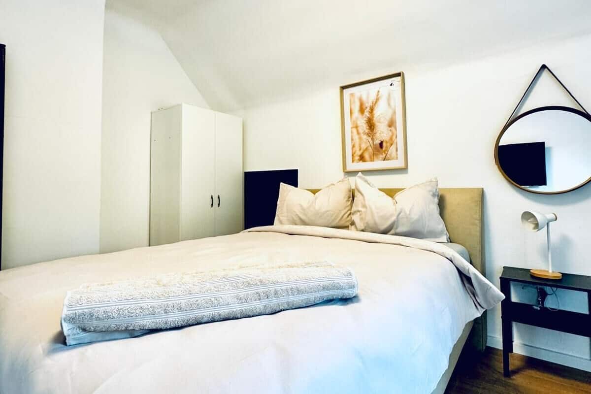 Un dormitorio con una cama grande, una mesita de noche con lámpara, un cuadro en la pared y una puerta al fondo.