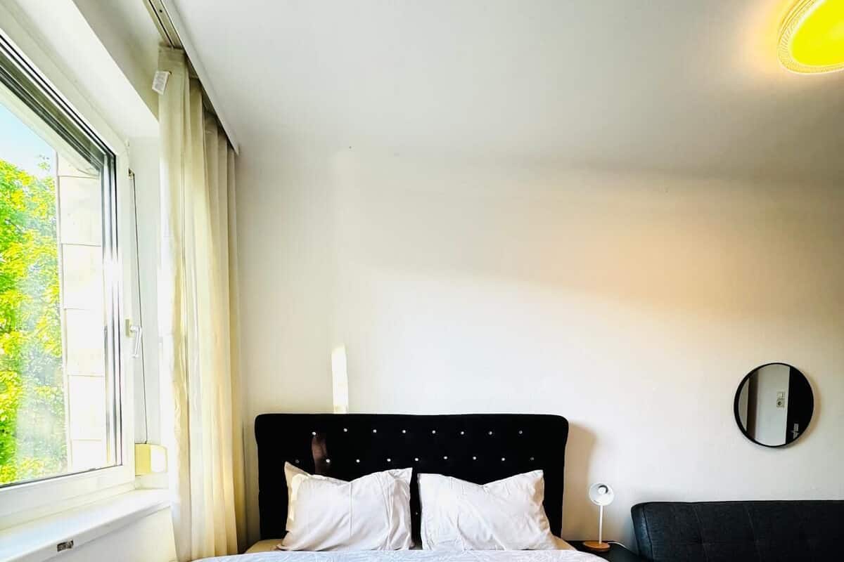 Una habitación de hotel con una cama grande, una mesita de noche con lámpara, un espejo redondo en la pared y una ventana con cortinas.