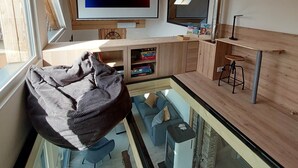 A modern living room with a wooden ceiling, a large TV displaying 'NETFLIX', a glass dining area, and a cozy seating area with a bean bag. - Le Dreispitz : Gîte d'Exception in Alsace (Gresswiller)
