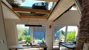 A modern kitchen with wooden flooring, a dining area with a table and benches, and large windows offering views of greenery and a blue-roofed building. - Le Dreispitz : Gîte d'Exception in Alsace (Gresswiller)