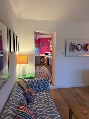 A living room with a patterned sofa, a lamp, a framed artwork, and a view into a kitchen with a pink wall and a clock.