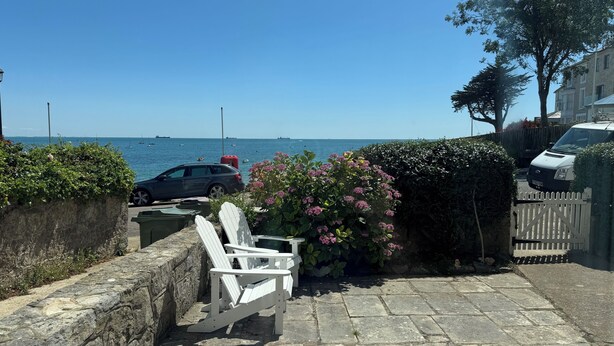 A stone-paved patio with a white Adirondack chair, a parked car, and a view of the sea with boats.
