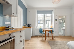 A modern kitchen with white cabinets, a wooden countertop, and a built-in oven. There is a dining area with a round table and blue chairs. - Melbourne Place (No 23) (St Andrews)