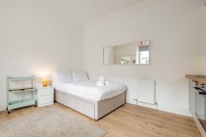 A bedroom with a bed, a nightstand with a lamp, a small shelf, a radiator, and a wooden floor. - Melbourne Place (No 23) (St Andrews)