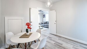 A modern dining area with a white table and chairs, a hanging light fixture, and a view into a living room with a sofa and a painting. - Furnished 1BR Near Downtown & UH – Monthly OK (Houston)