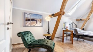 A room with a green upholstered chair, a small wooden table, a bed, and a painting of a beach scene.