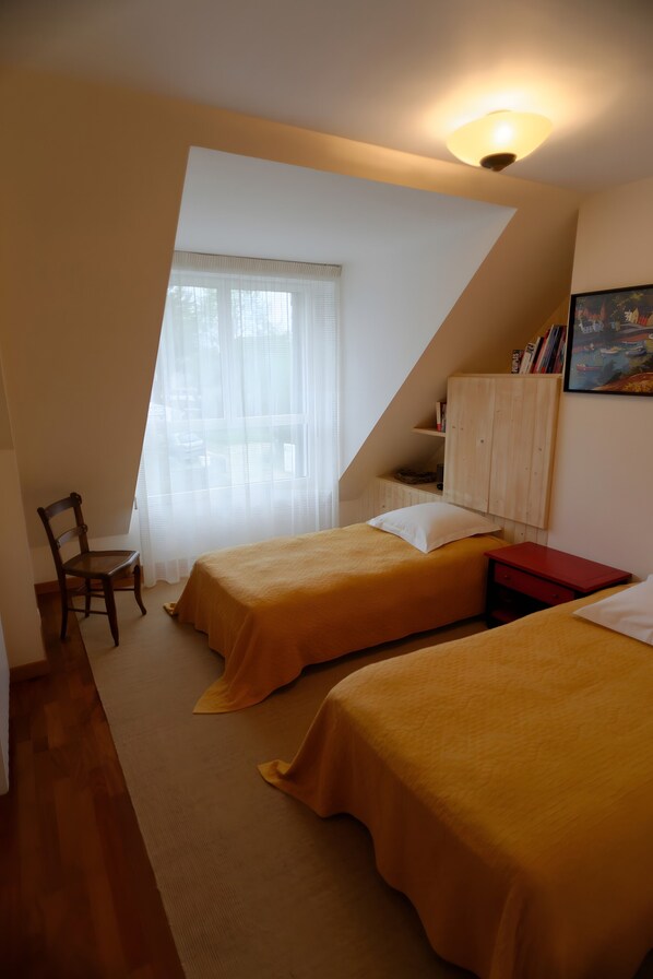 A bedroom with two beds, a chair, a desk, a shelf with books, and a painting on the wall.