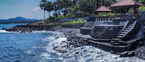 Una zona costera con una escalera de piedra que conduce a un mirador junto al mar, rodeada de palmeras y un cielo despejado.