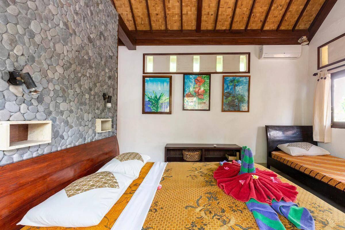 Ein Schlafzimmer mit Steinwand, Holzbalkendecke und einem Bett mit gemustertem Überwurf. An der Wand hängen drei Gemälde und es gibt ein kleines Regal.