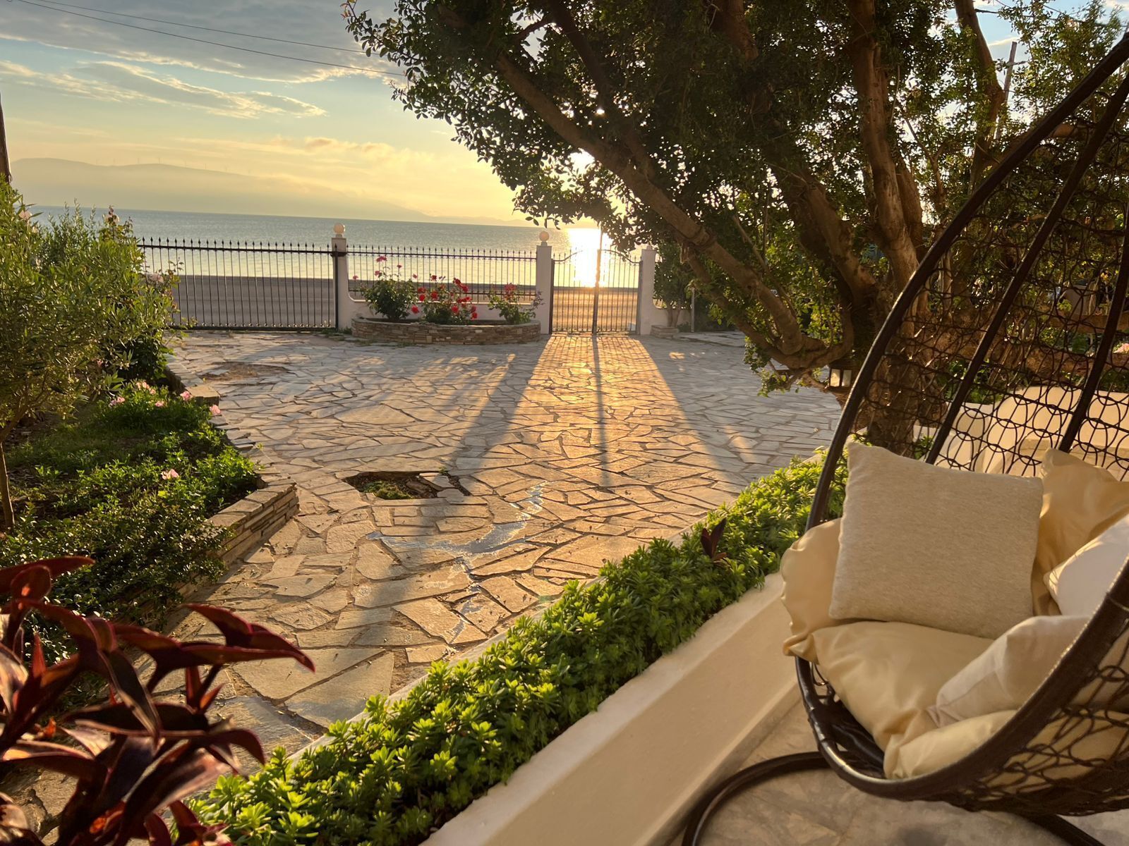A stone-paved patio with a hanging chair and cushions, overlooking a body of water and a fence.