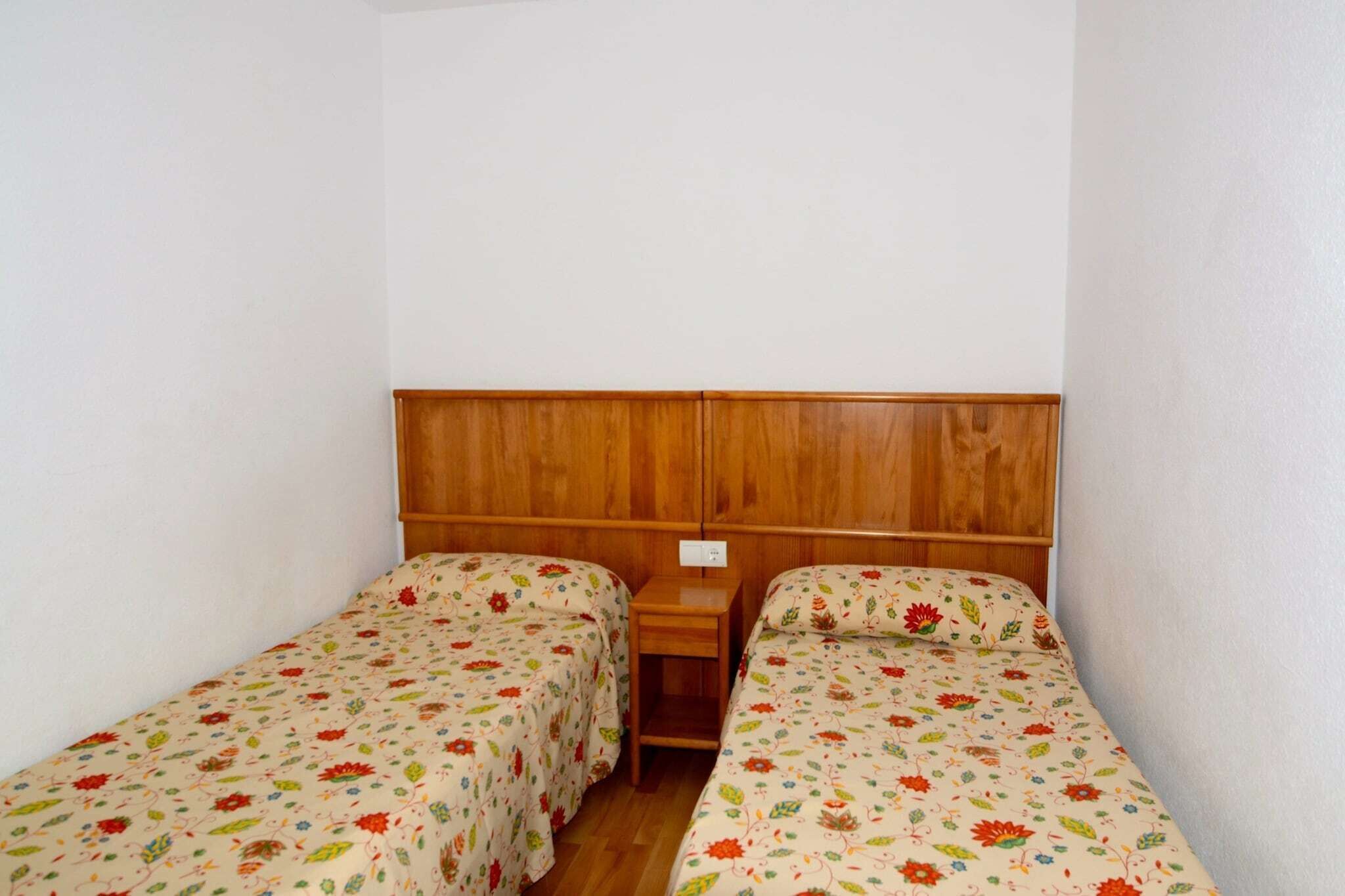 Ein kleines, schlichtes Zimmer mit zwei Einzelbetten, einem Holzkopfteil und einem Nachttisch dazwischen.