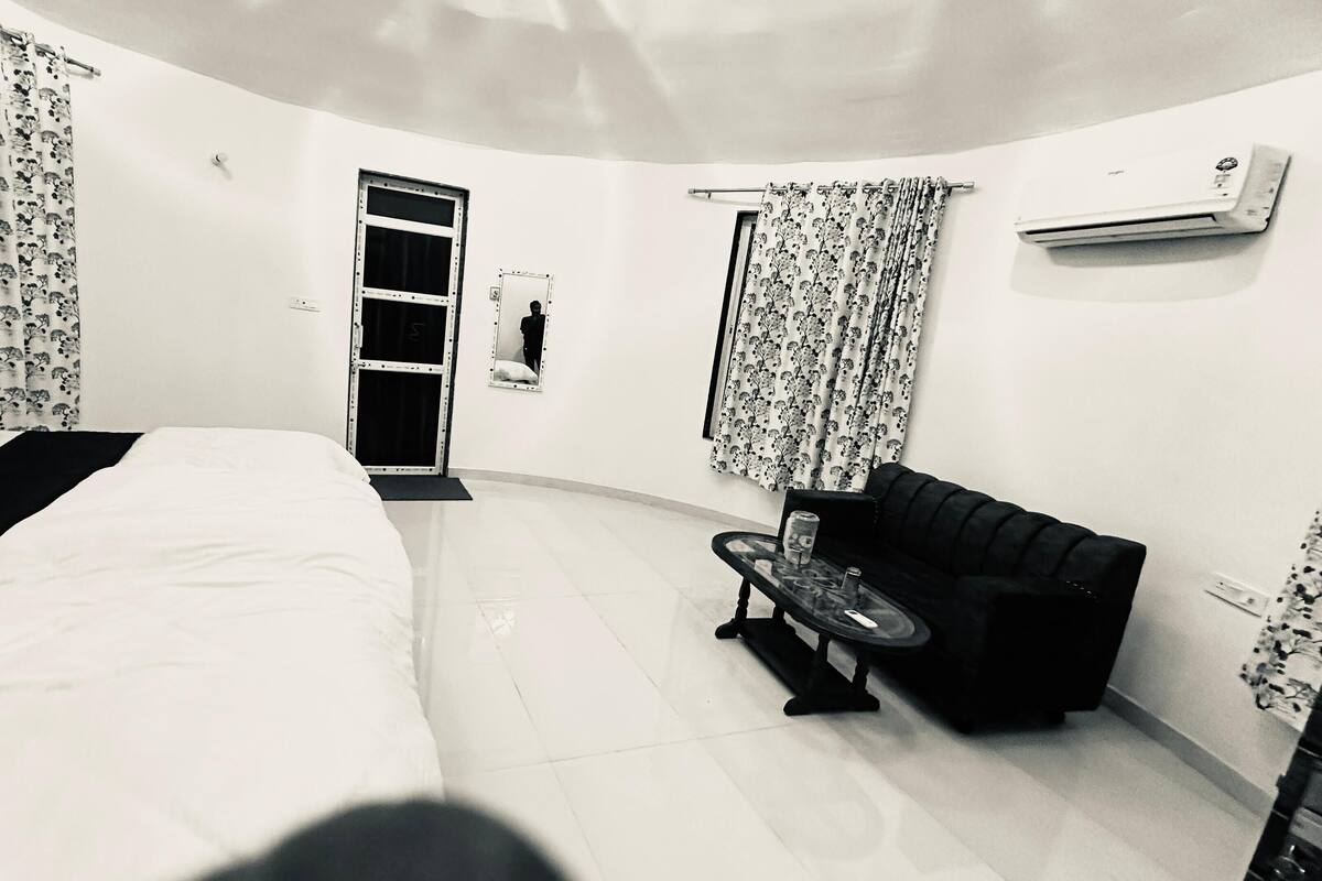 Habitación de hotel con cama, un pequeño sofá, una mesa, un espejo, una puerta y un equipo de aire acondicionado.