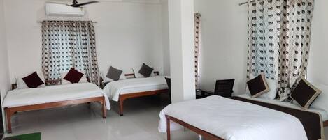 A hotel room with two single beds, a desk, and a chair. There is a ceiling fan, an air conditioning unit, and patterned curtains.