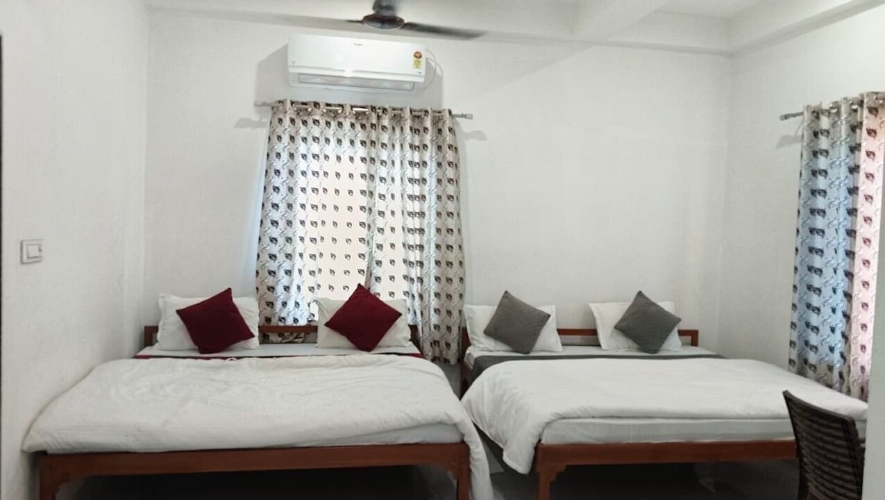 A room with two single beds, a ceiling fan, an air conditioning unit, and patterned curtains.