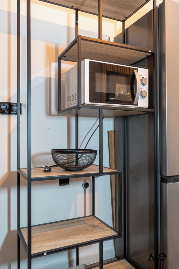 Une cuisine avec un four à micro-ondes posé sur une étagère, un saladier noir en treillis métallique et une planche à découper en bois.