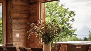 Ein Holzespeisetisch mit sechs Stühlen, eine Vase mit Blumen und Blick auf Bäume und Berge durch große Fenster.