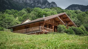 Eine Holzhütte mit Balkon, umgeben von üppiger Vegetation und Bergen im Hintergrund.