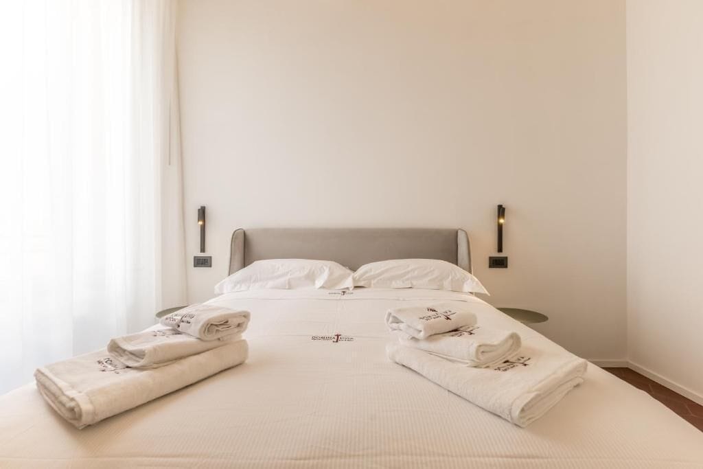 Un lit bien fait, avec du linge de lit et des serviettes blancs, une tête de lit grise et deux appliques murales.