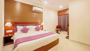 A hotel room with a large bed, two bedside lamps, an air conditioning unit, a ceiling fan, a small table with a chair, and a window with curtains. - Classio Inn Hotel (Bengaluru)