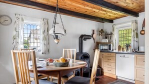 A dining area with a wooden table, chairs, a hanging light fixture, a wood stove, and a kitchen with a microwave and cabinets.