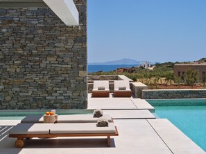 A poolside area with lounge chairs and a stone wall, overlooking a clear blue sea and a distant mountain. - Luxury Villa Serenely (Drios)