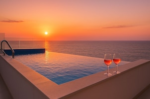 Una piscina infinita con barandilla de vidrio, dos copas de vino tinto y vistas al atardecer sobre el océano.