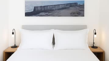 A neatly made bed with white linens, a large framed picture of a coastal scene above it, and two bedside lamps on wooden nightstands.