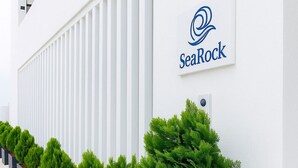 A modern building with a white facade, a SeaRock logo, and a row of neatly arranged potted evergreen trees. - SeaRock Suites (Aamchit)