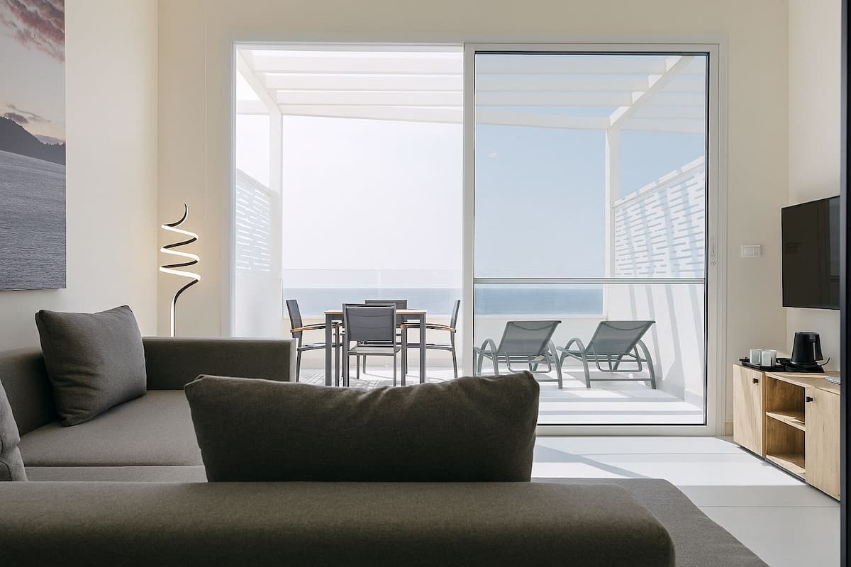 A modern living room with a sofa, a coffee table, and a TV. There is a balcony with a table and chairs, and a view of the sea.
