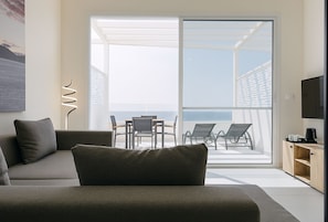 Una sala de estar moderna con un sofá, una mesa de centro y un televisor. Hay un balcón con una mesa y sillas, y vista al mar.