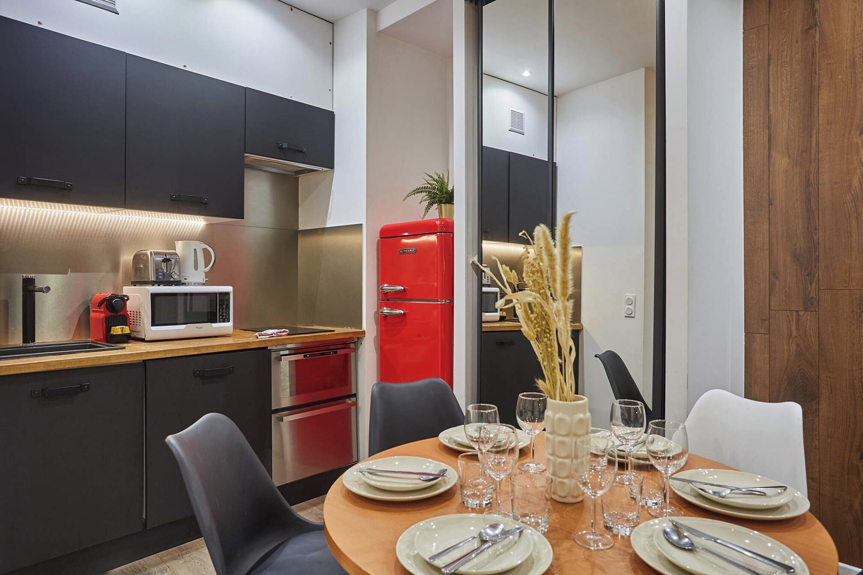Une cuisine moderne avec des meubles de couleur sombre, un réfrigérateur rouge et un coin repas avec une table en bois, mise pour quatre personnes.