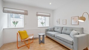 Een moderne woonkamer met een grijze bank, een kleine oranje fauteuil, een rond bijzettafeltje en twee ramen met zonwering.