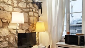 Une petite pièce agréable, avec un mur en pierre, un bureau blanc, une télévision, une lampe, une chaise, un radiateur et une fenêtre avec des rideaux.