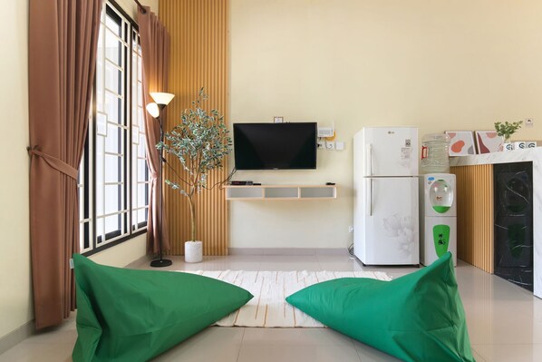 Una sala de estar moderna con un televisor de pantalla plana, un refrigerador, una planta en maceta y cojines verdes.