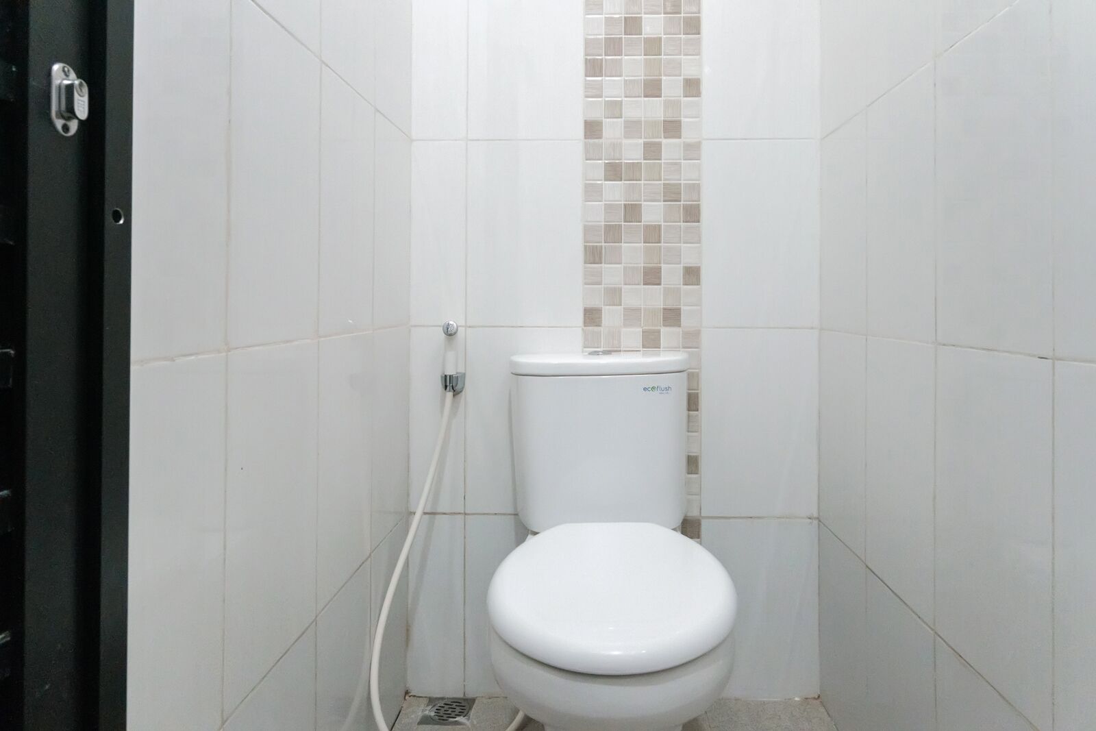 Un baño pequeño con inodoro blanco, pared con mosaicos color beige y zona de ducha blanca, y una puerta con manija plateada.