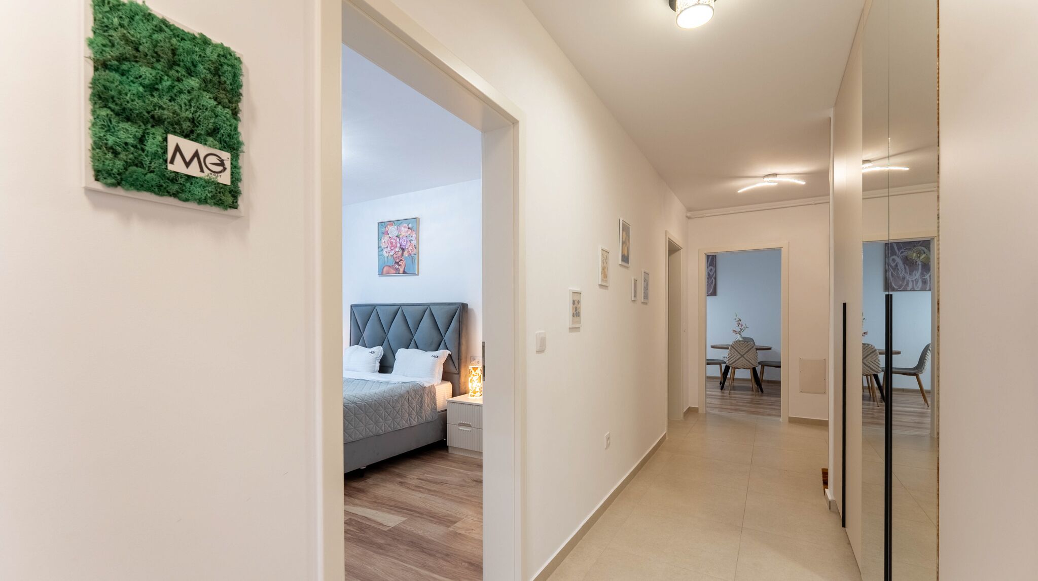 A modern hotel corridor with a bed, a green wall with a logo, and a dining area with chairs and a table.