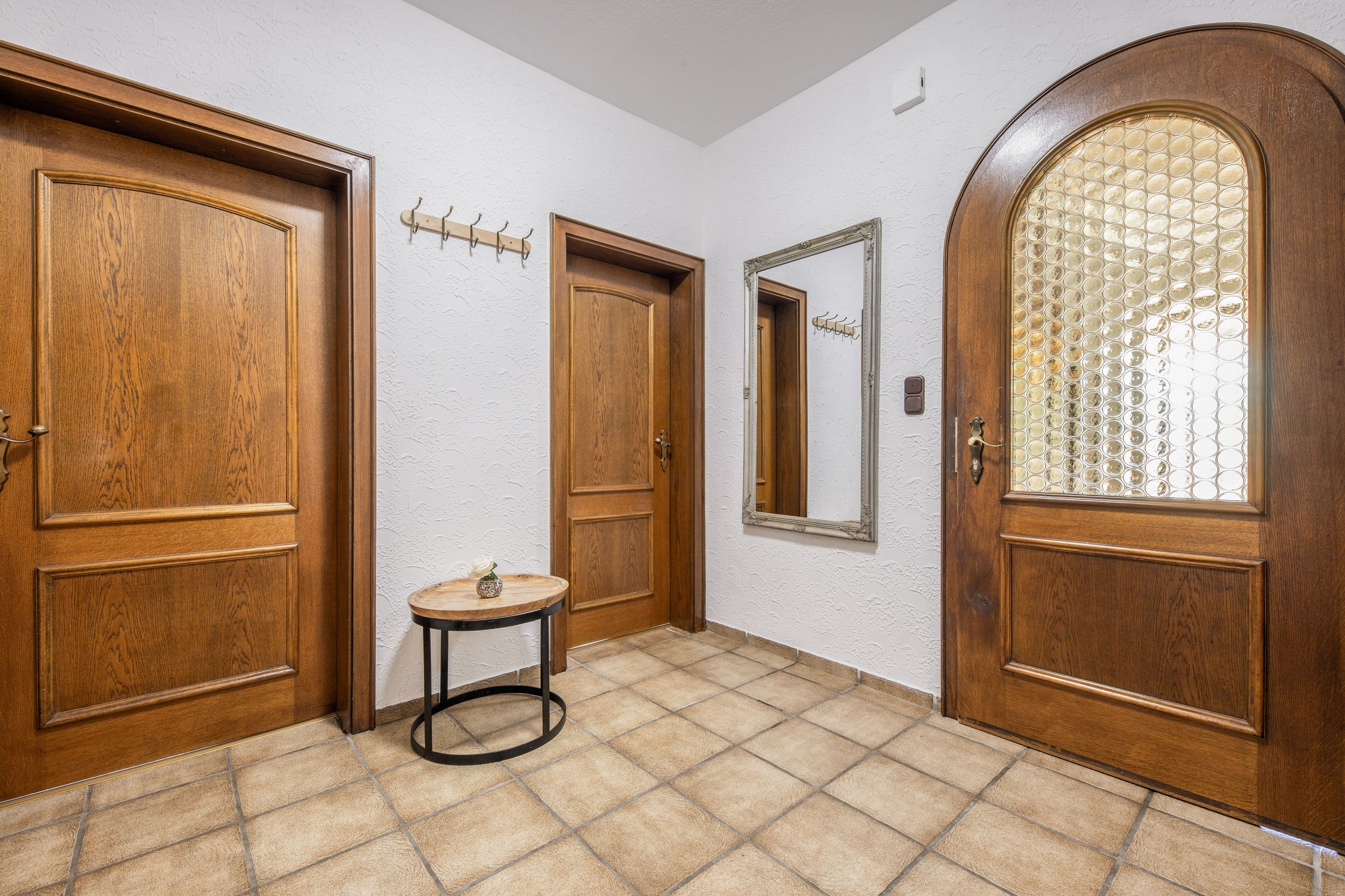 Un couloir avec des portes en bois, une petite table ronde surmontée d’un vase et un miroir accroché au mur.