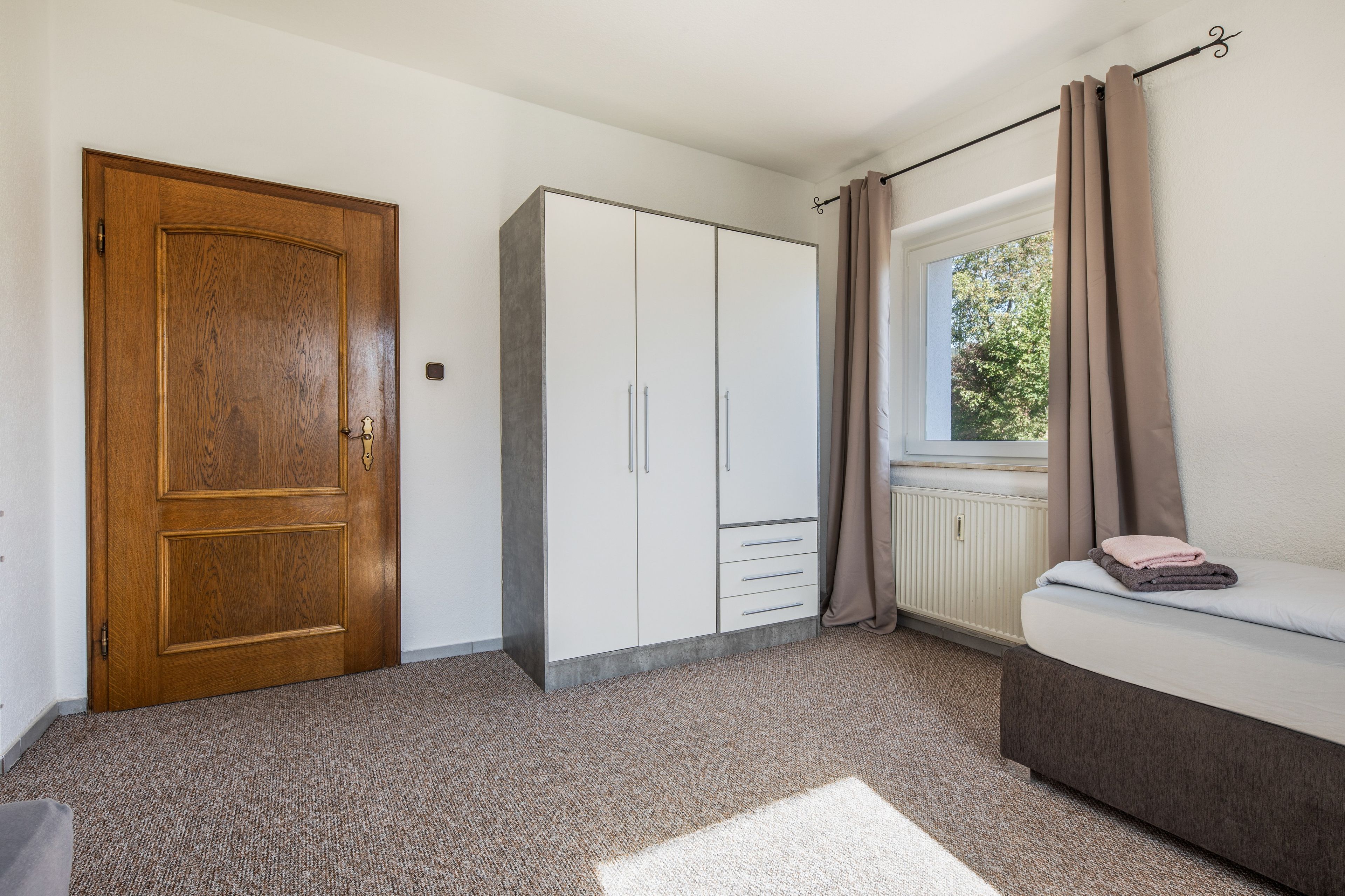 Ein Schlafzimmer mit Holztür, einem Kleiderschrank, einem Heizkörper, einem Fenster mit Vorhängen und einem Bett mit Handtüchern.