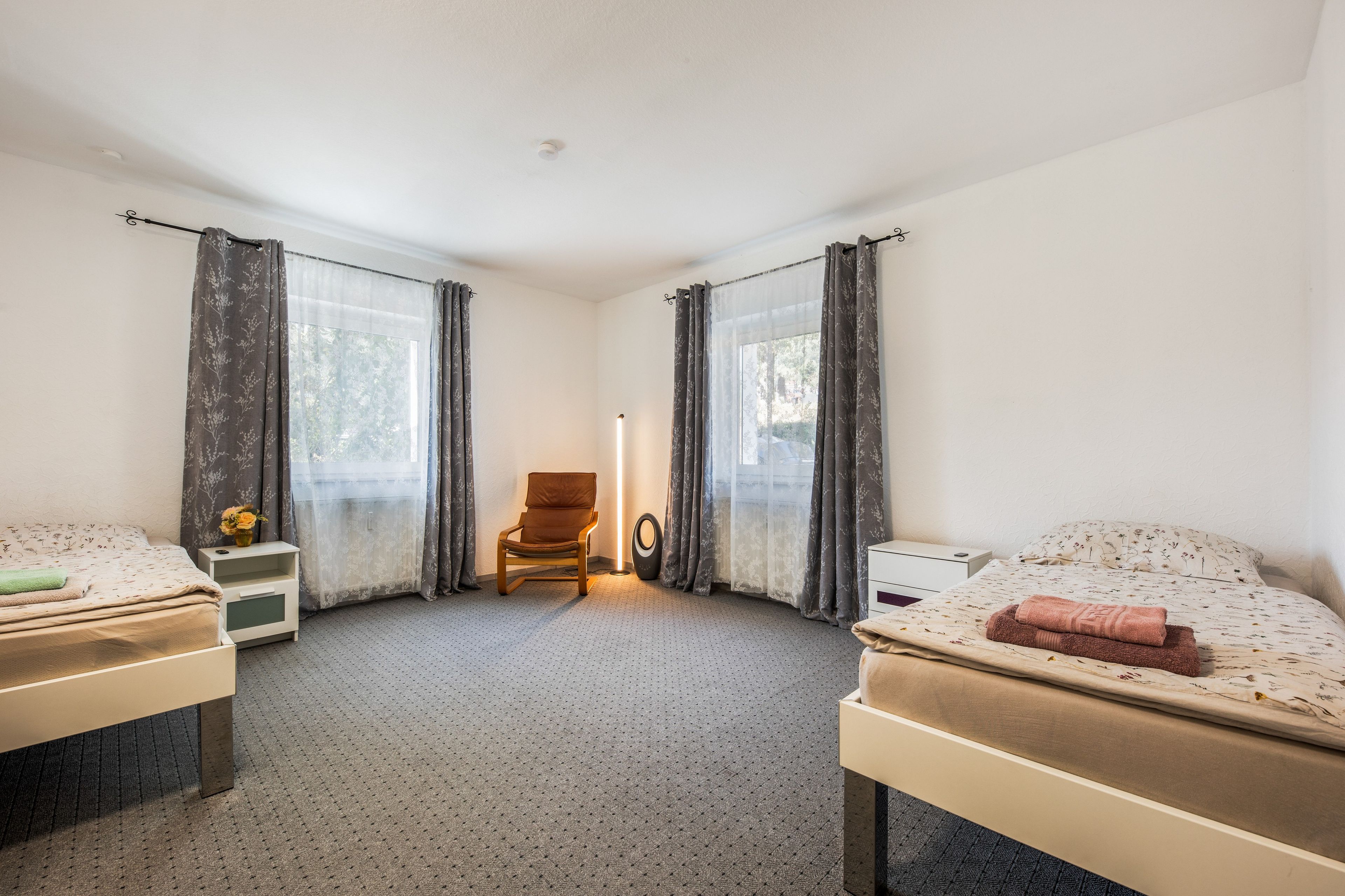 Ein Zimmer mit zwei Betten, einem Sessel, einem Nachttisch, einer Lampe und einem fensterbepflanzten Fenster.