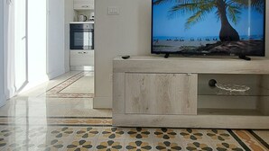 A modern living room with a flat-screen TV, a kitchen in the background, and patterned flooring. - Sea Vibes Rome - Relax by the sea and reach Rome in minutes (Lido di Ostia)