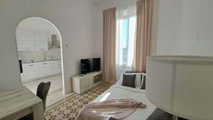 A room with a TV on a wooden cabinet, a bed with a white mattress and a pink blanket, a small table with a chair, and a kitchen area with cabinets and a sink. - Sea Vibes Rome - Relax by the sea and reach Rome in minutes (Lido di Ostia)