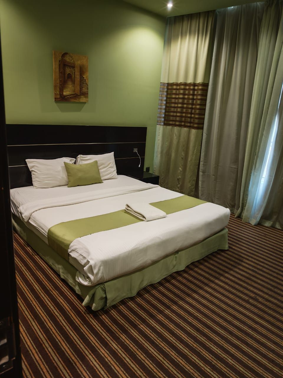 A hotel room with a large bed, a headboard, a painting on the wall, and curtains.
